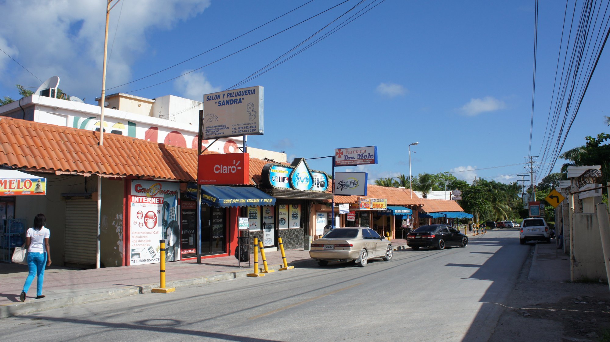 Photographie d'une rue commerçante de Bávaro, Punta Cana : une arcade aux tuiles rouges abritant de petits commerces aux enseignes peintes à la main, des voitures garées en épi, des câbles tendus entre les poteaux — le visage quotidien du secteur où les propriétaires de River Island se rendent en cinq minutes.